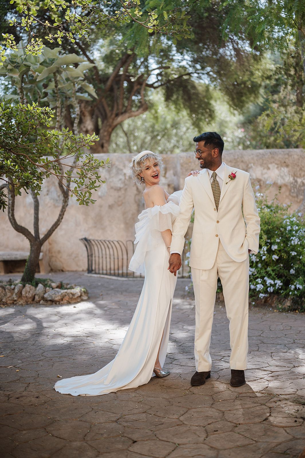Bride in white off-shoulder gown and groom in beige suit hold hands smiling in a sunlit courtyard.