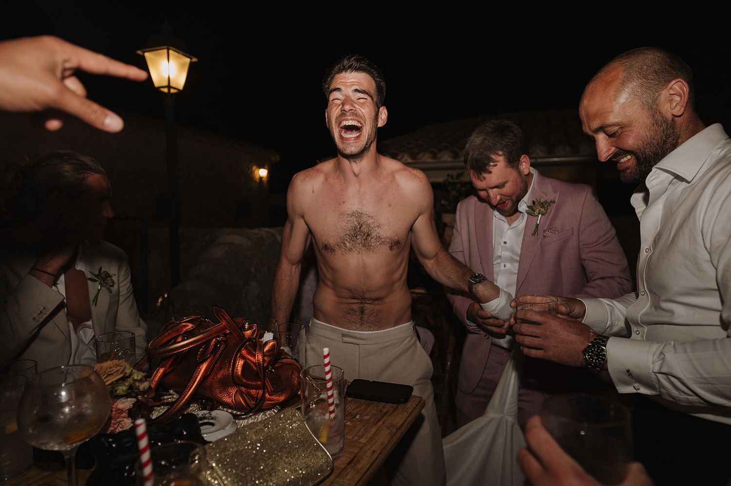 Shirtless man laughing joyfully with guests in suits at an outdoor evening wedding party.