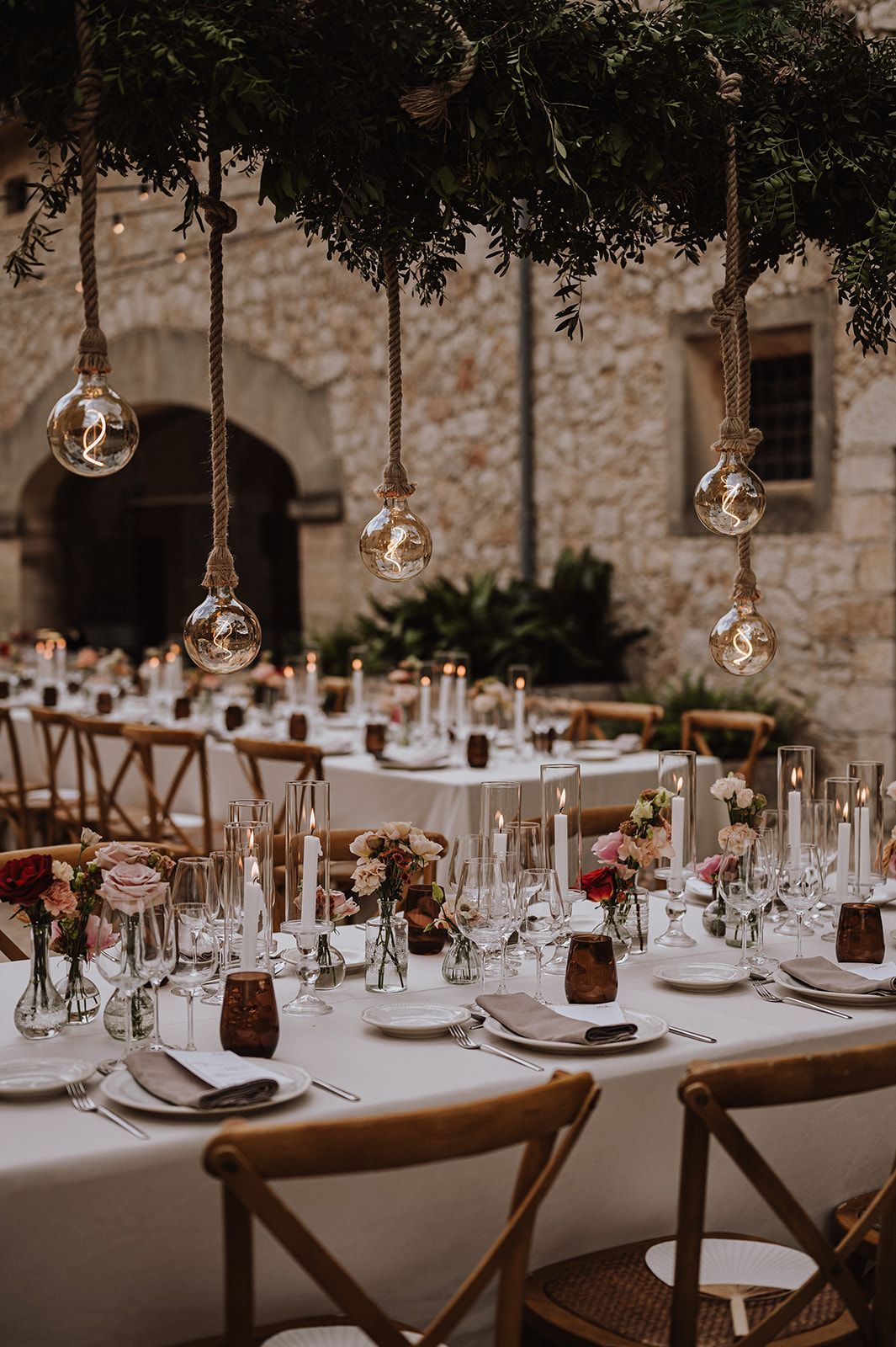 Wedding reception table set with white linens, crystal glassware, pink florals, and hanging Edison bulb lights.