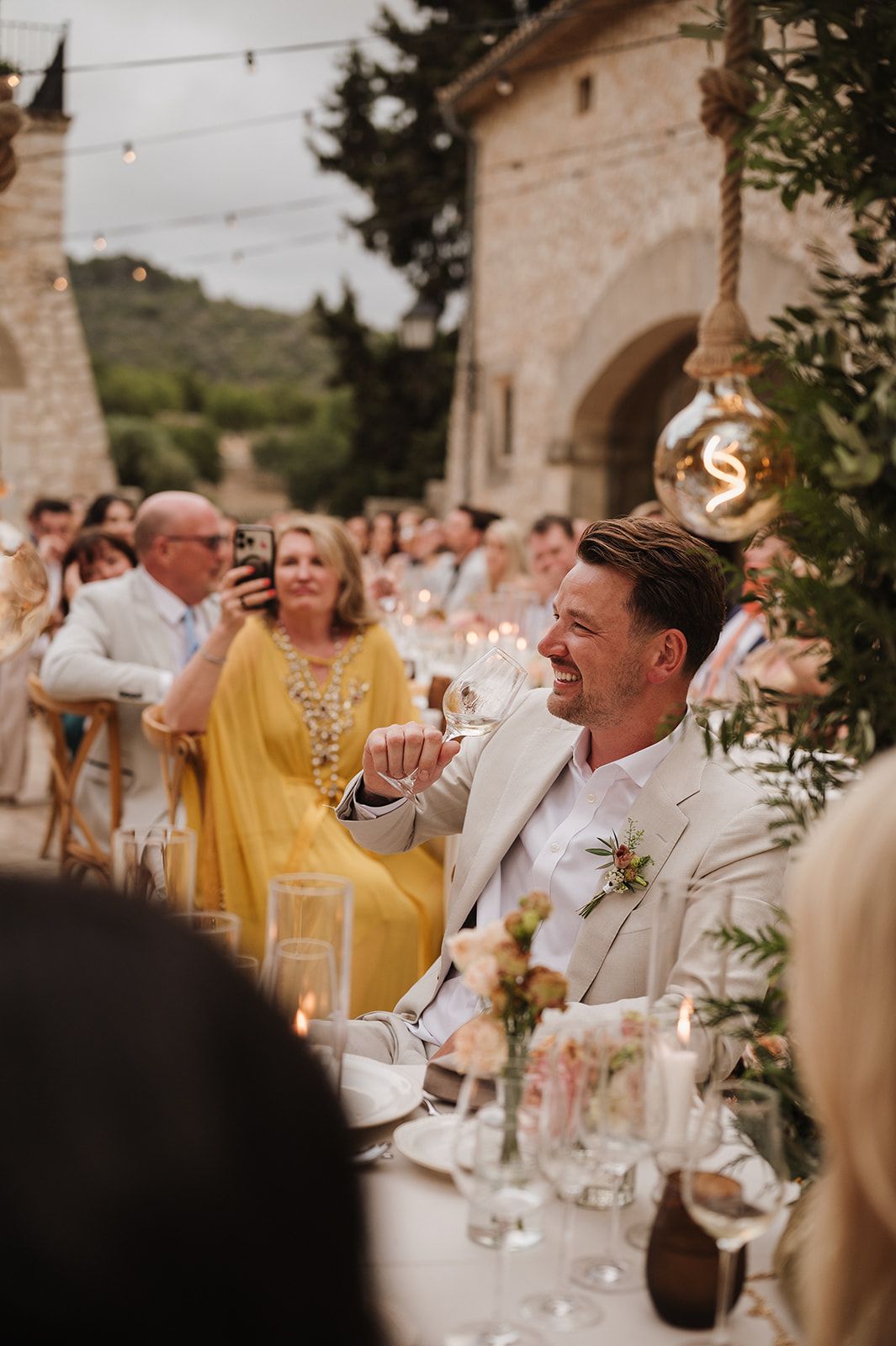 Smiling groom in cream suit applauding at outdoor wedding reception surrounded by guests and candles.