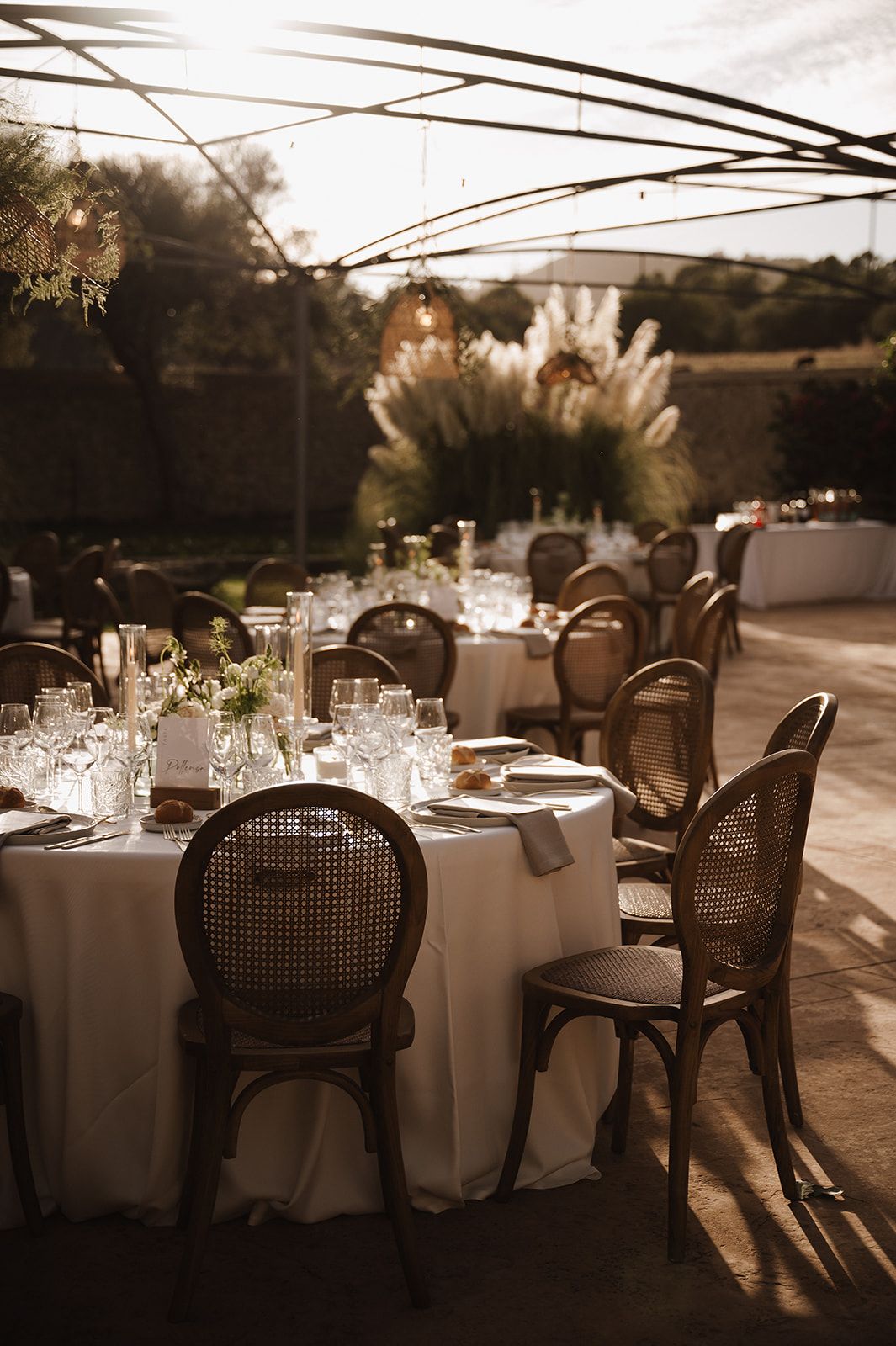 Elegant outdoor wedding reception with rattan chairs, white tablecloths, and pampas grass centerpieces.