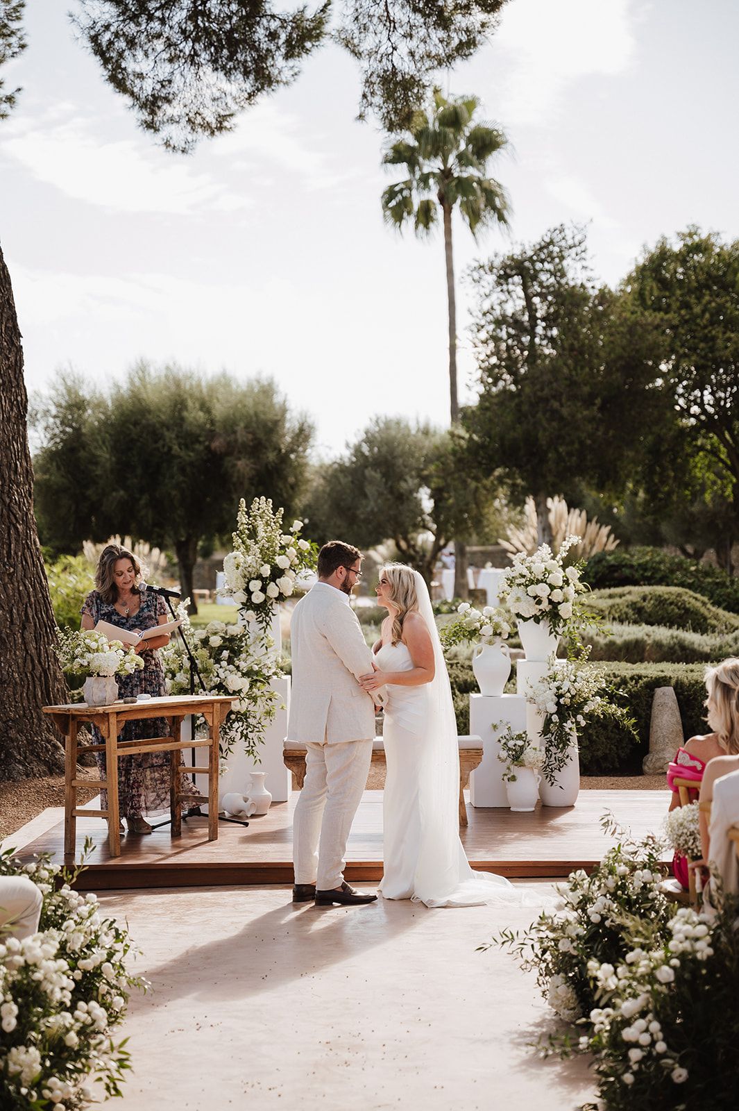 Couple exchanges vows at an elegant outdoor wedding ceremony surrounded by white floral arrangements and lush greenery.