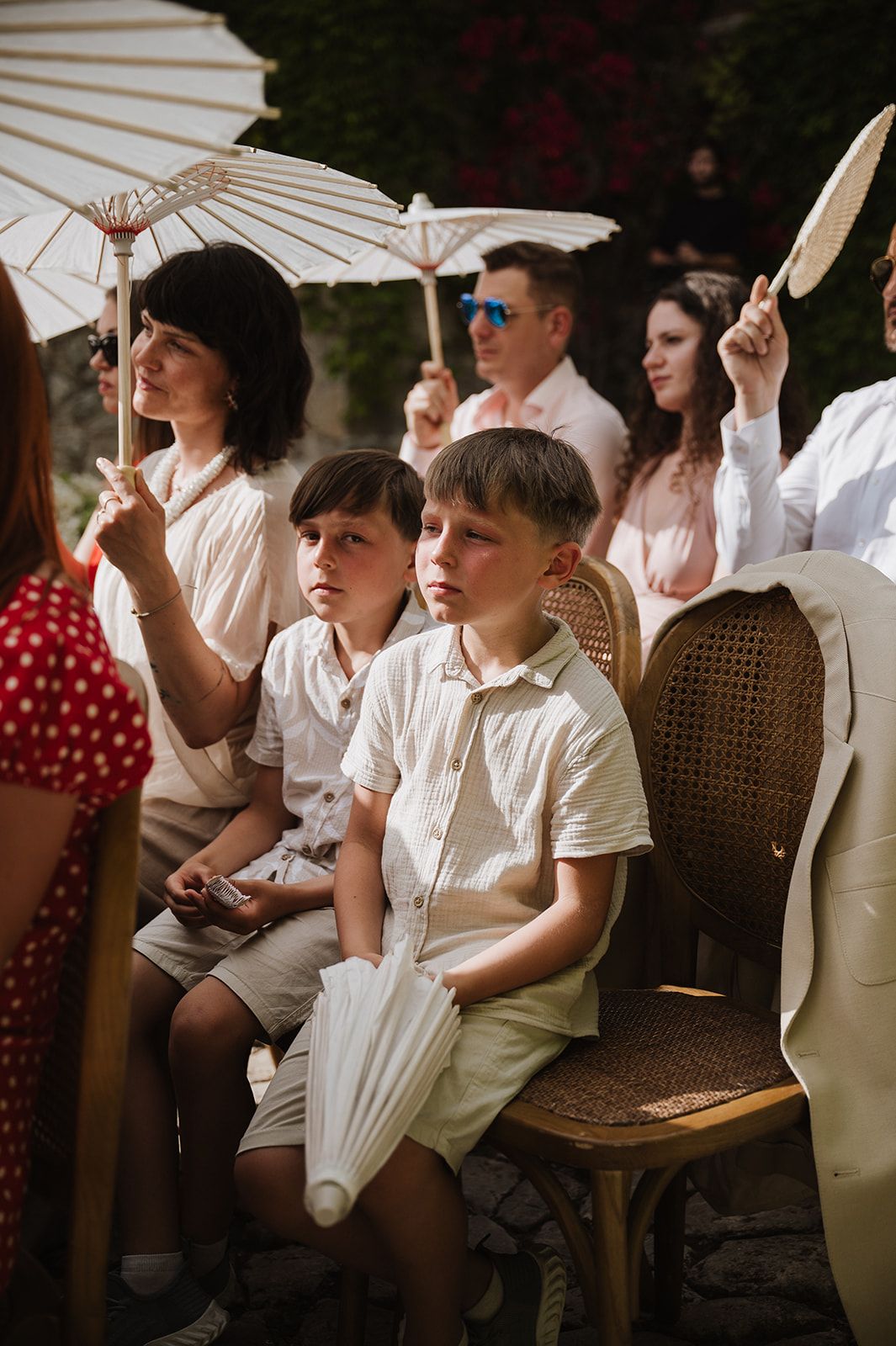Young boys in white outfits sit among wedding guests holding parasols during an outdoor ceremony.