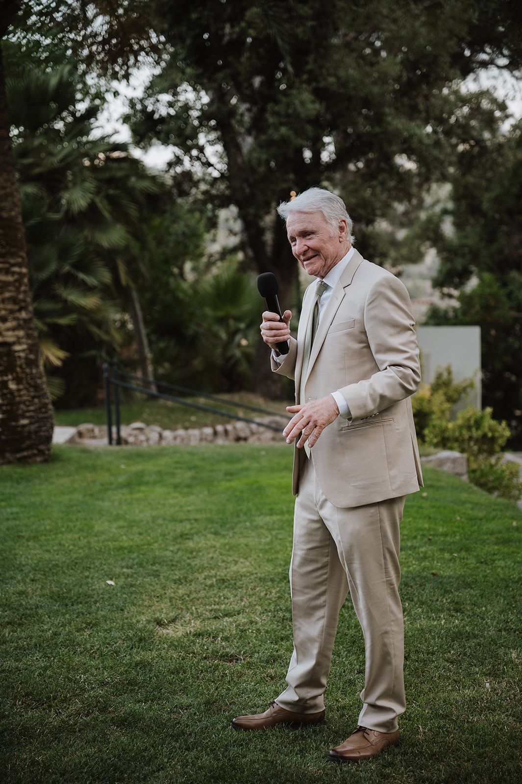 Elegant older gentleman in beige suit delivering a wedding speech on green lawn surrounded by lush trees.