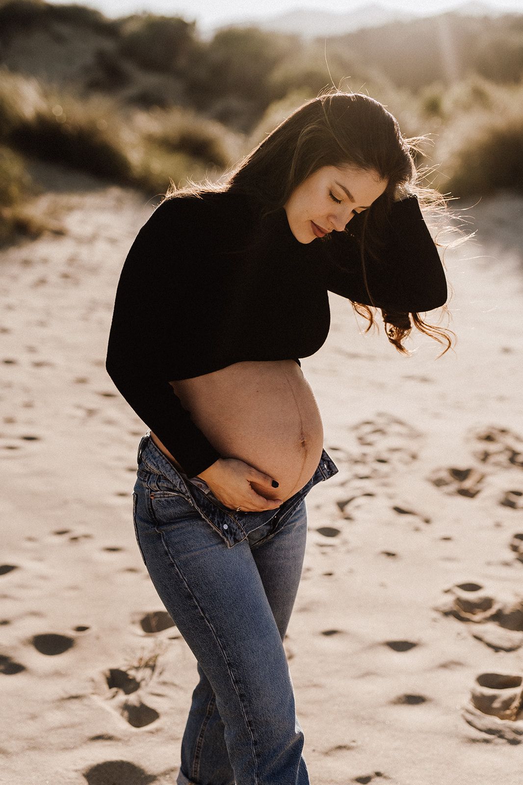 Pregnant woman in black crop top and jeans cradling her baby bump on a sandy beach at golden hour.