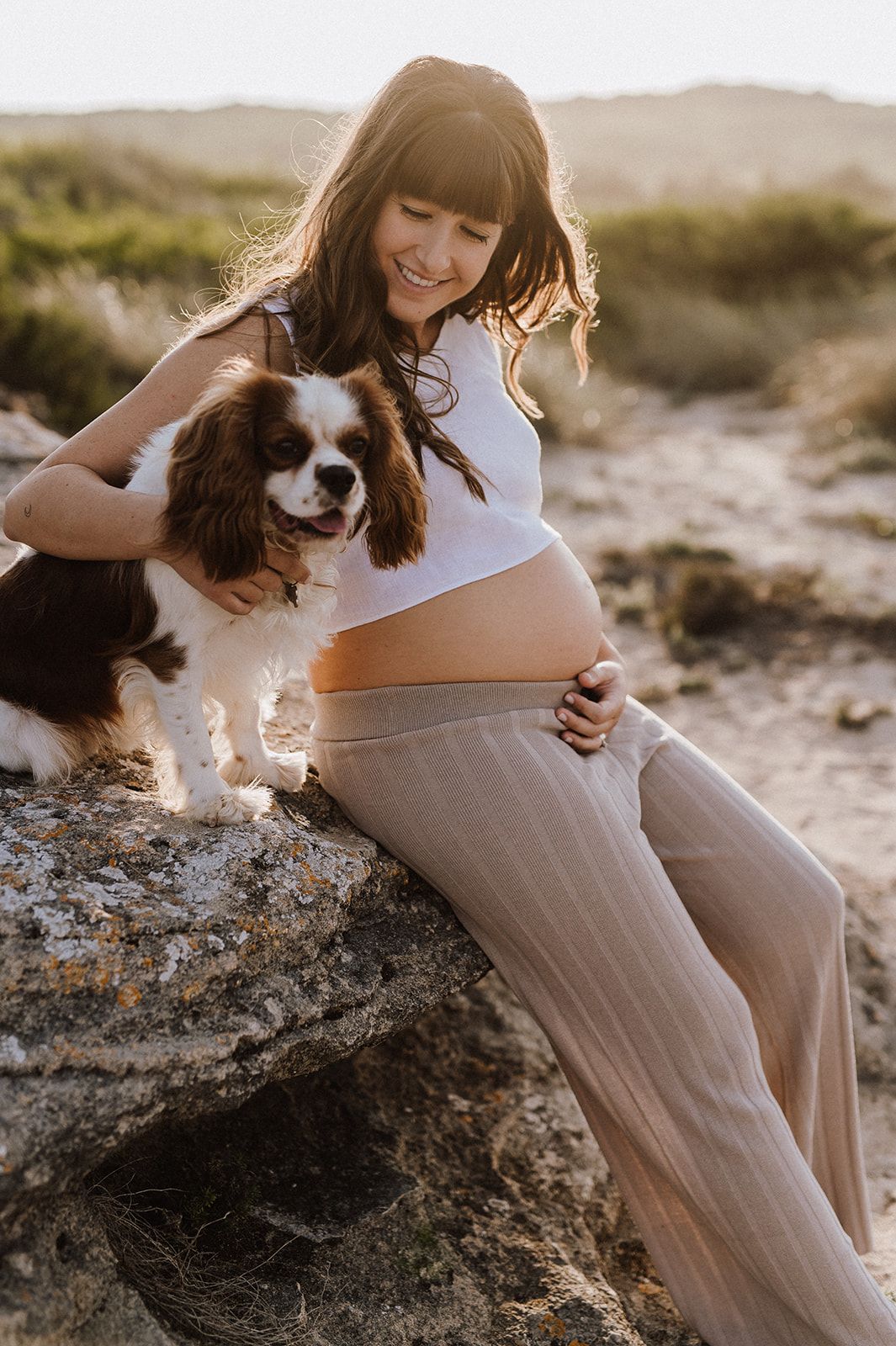 Pregnant woman in white top and sheer pants sits on rock with Cavalier King Charles Spaniel during golden hour outdoors.