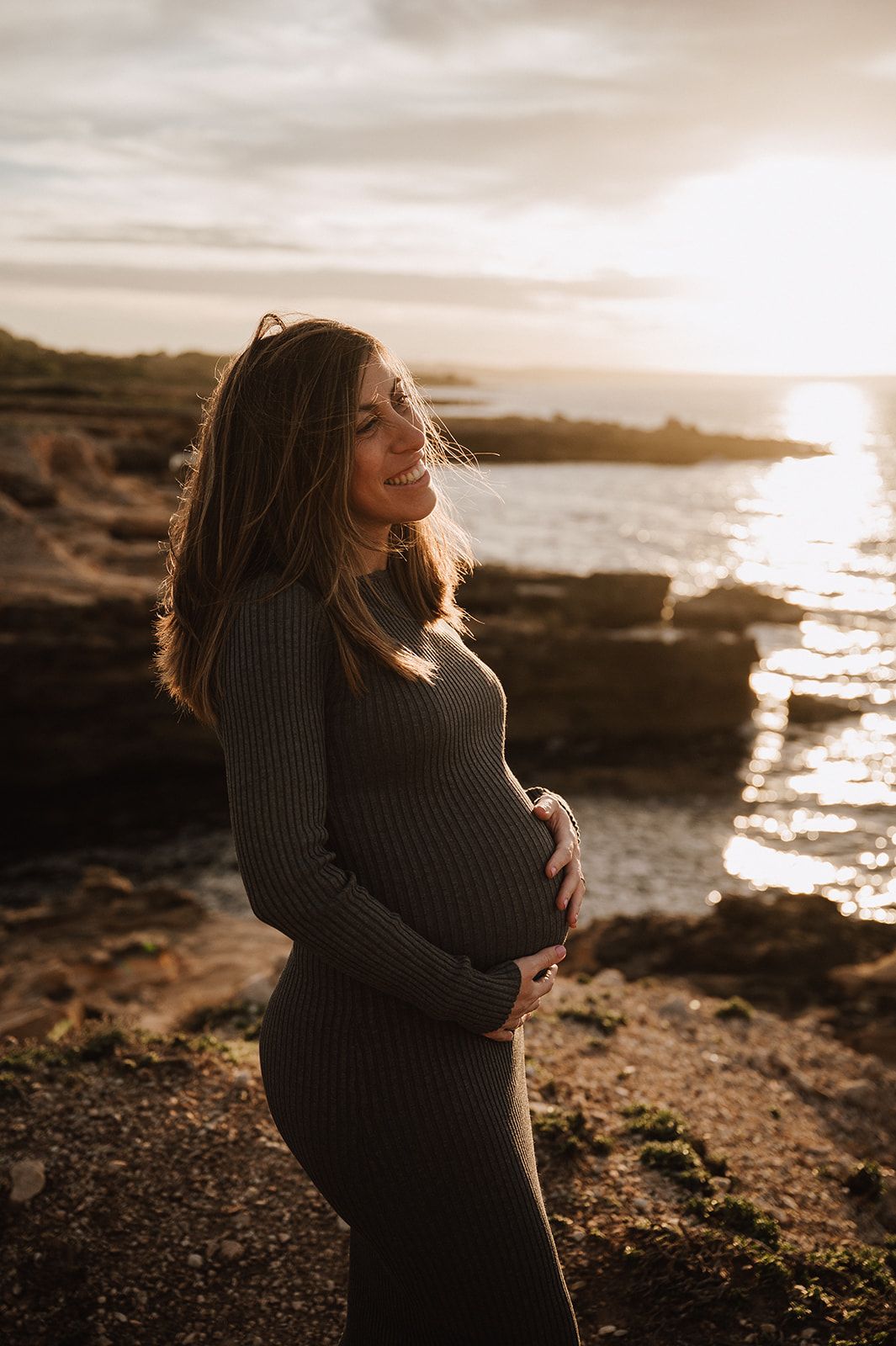 Smiling pregnant woman in gray ribbed dress cradling baby bump at golden hour by rocky ocean shore.