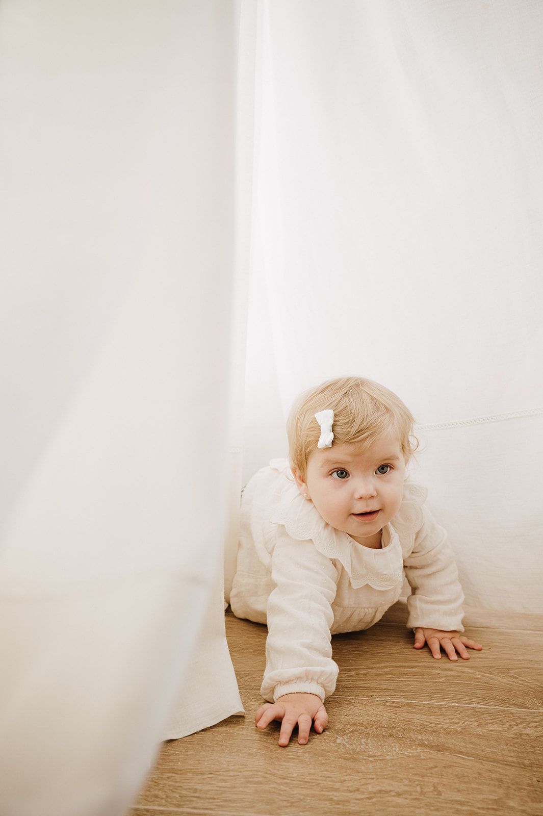 Baby in white outfit with bow clip crawling on hardwood floor near curtain.