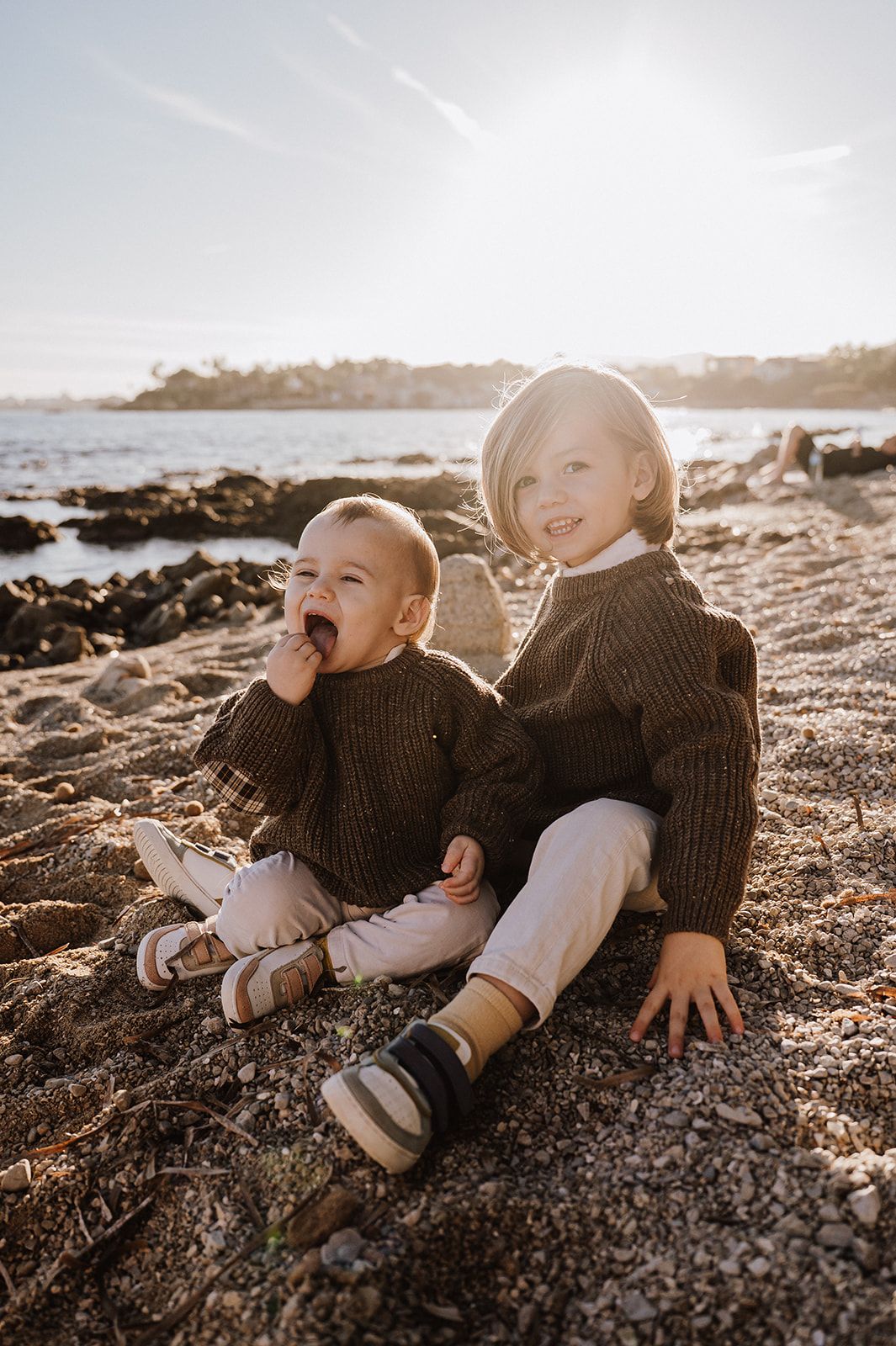 Two young siblings in matching brown knit sweaters sitting together on a sunny beach.