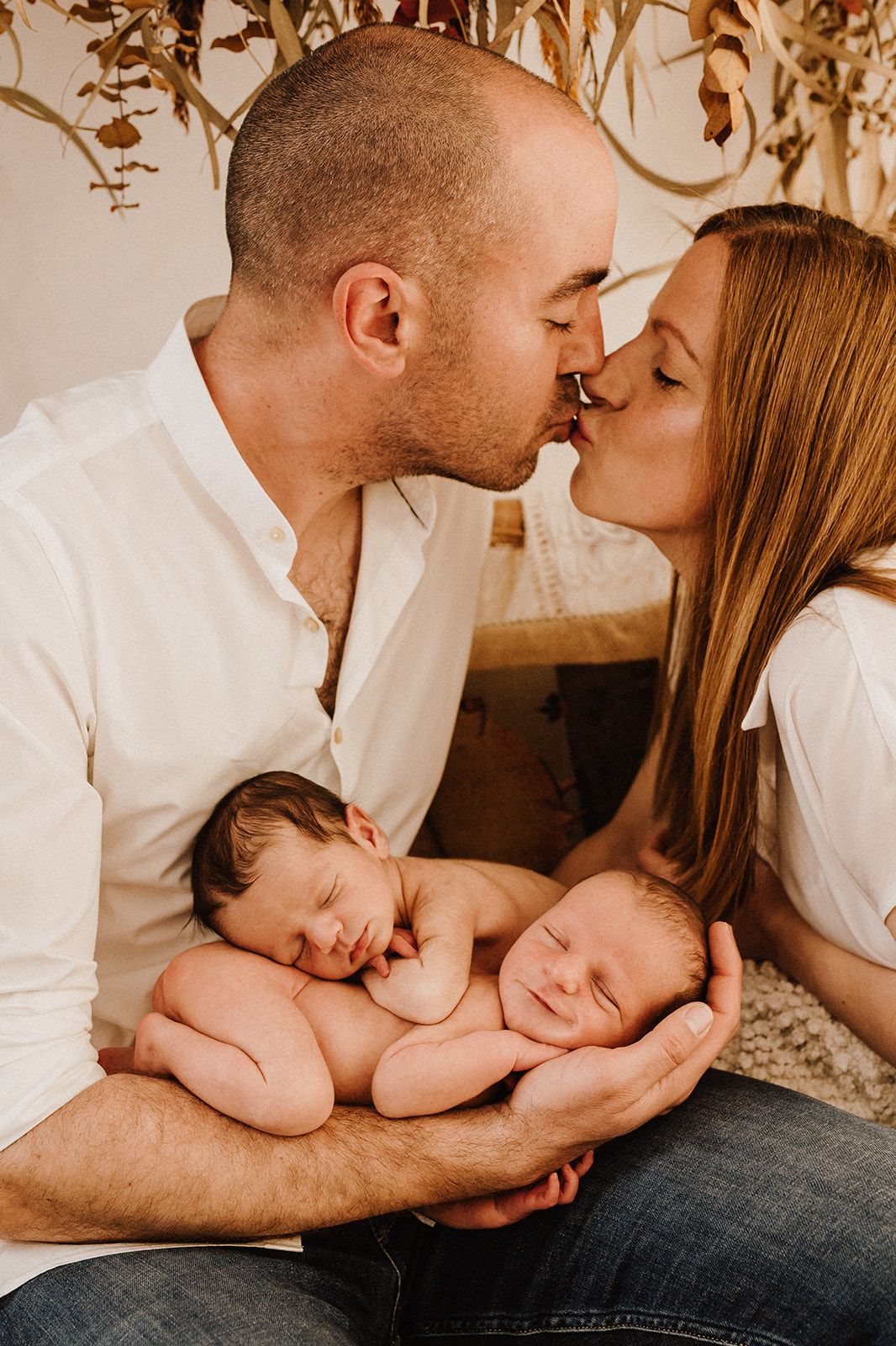 Parents kissing while holding their sleeping newborn twins in a warm, intimate family portrait photo.