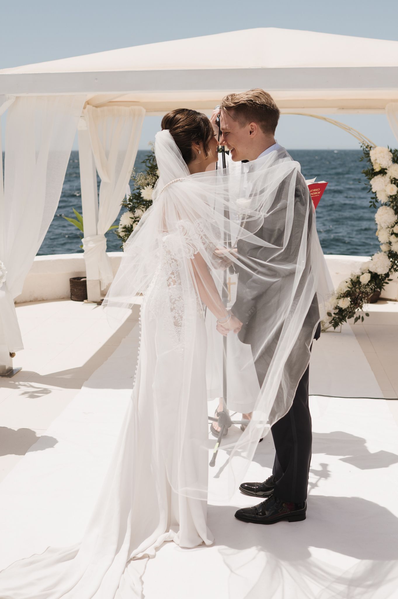Bride and groom sharing an intimate moment under a white floral arch by the ocean.