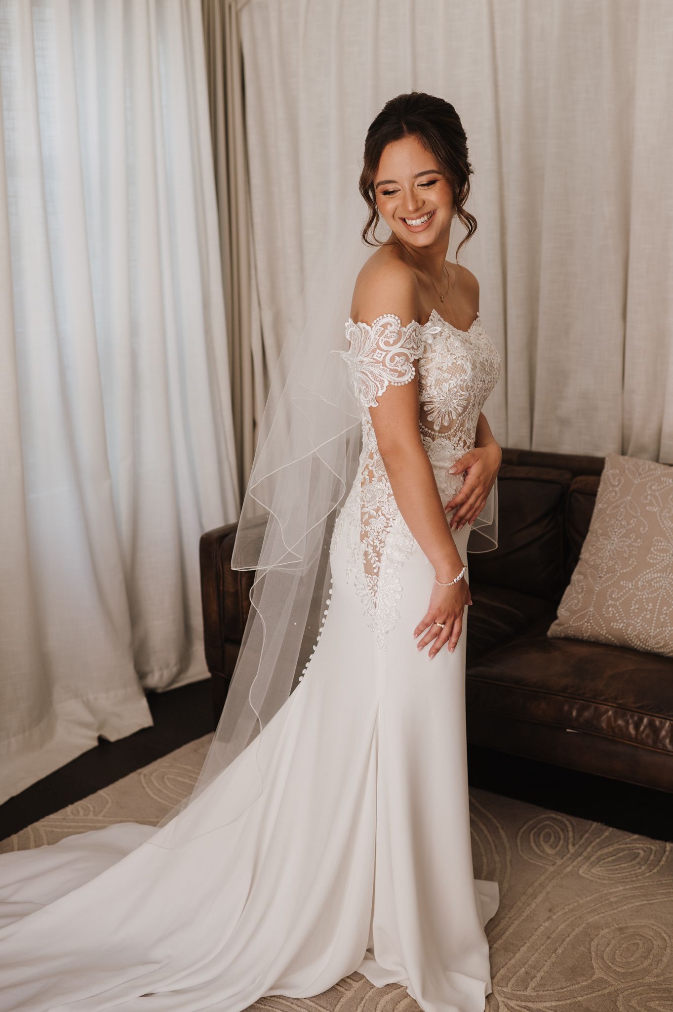 Smiling bride in off-shoulder lace wedding gown with veil and train poses in elegant hotel room.