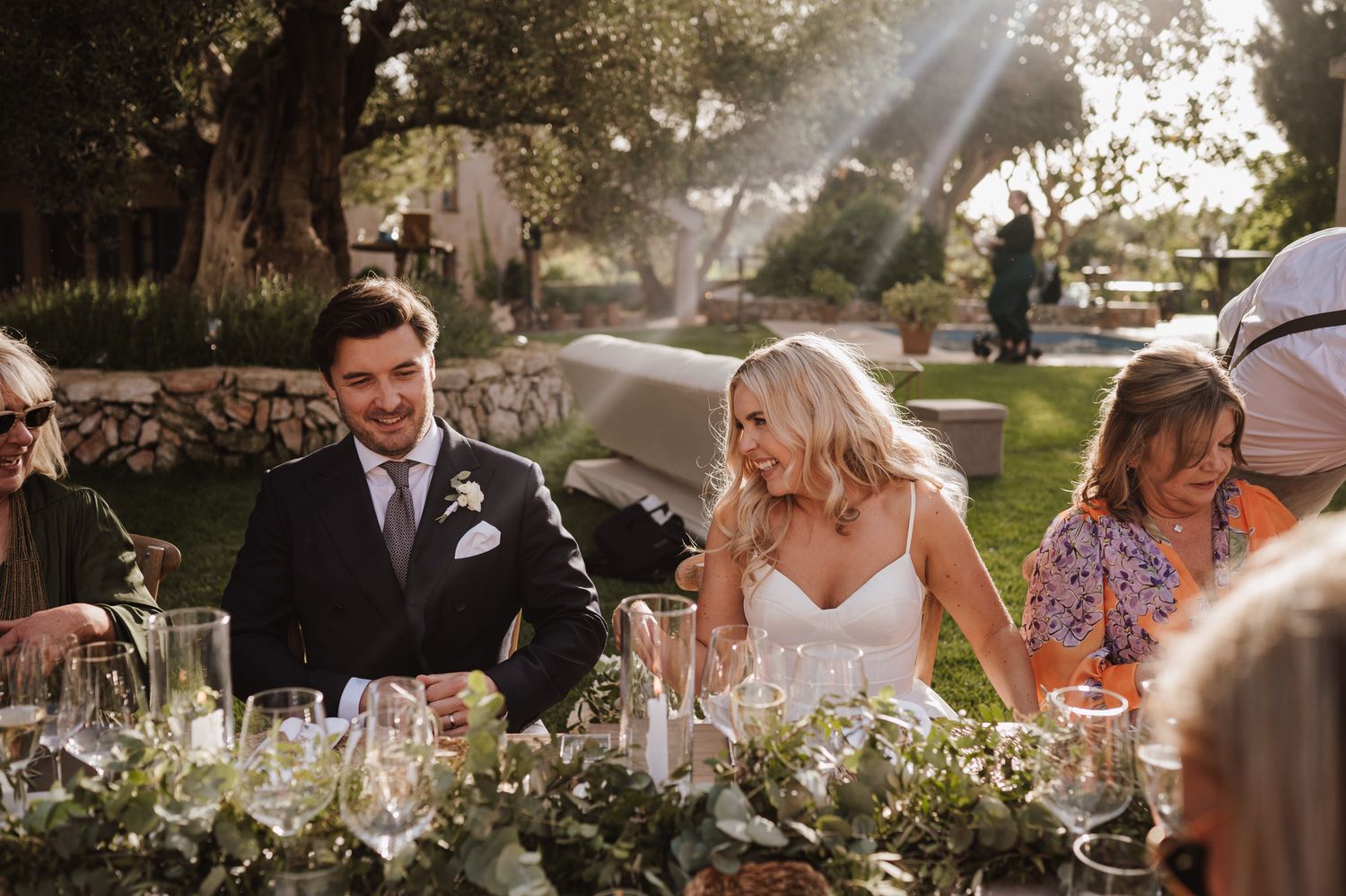 Bride and groom laugh together at a long outdoor wedding reception table adorned with lush greenery.