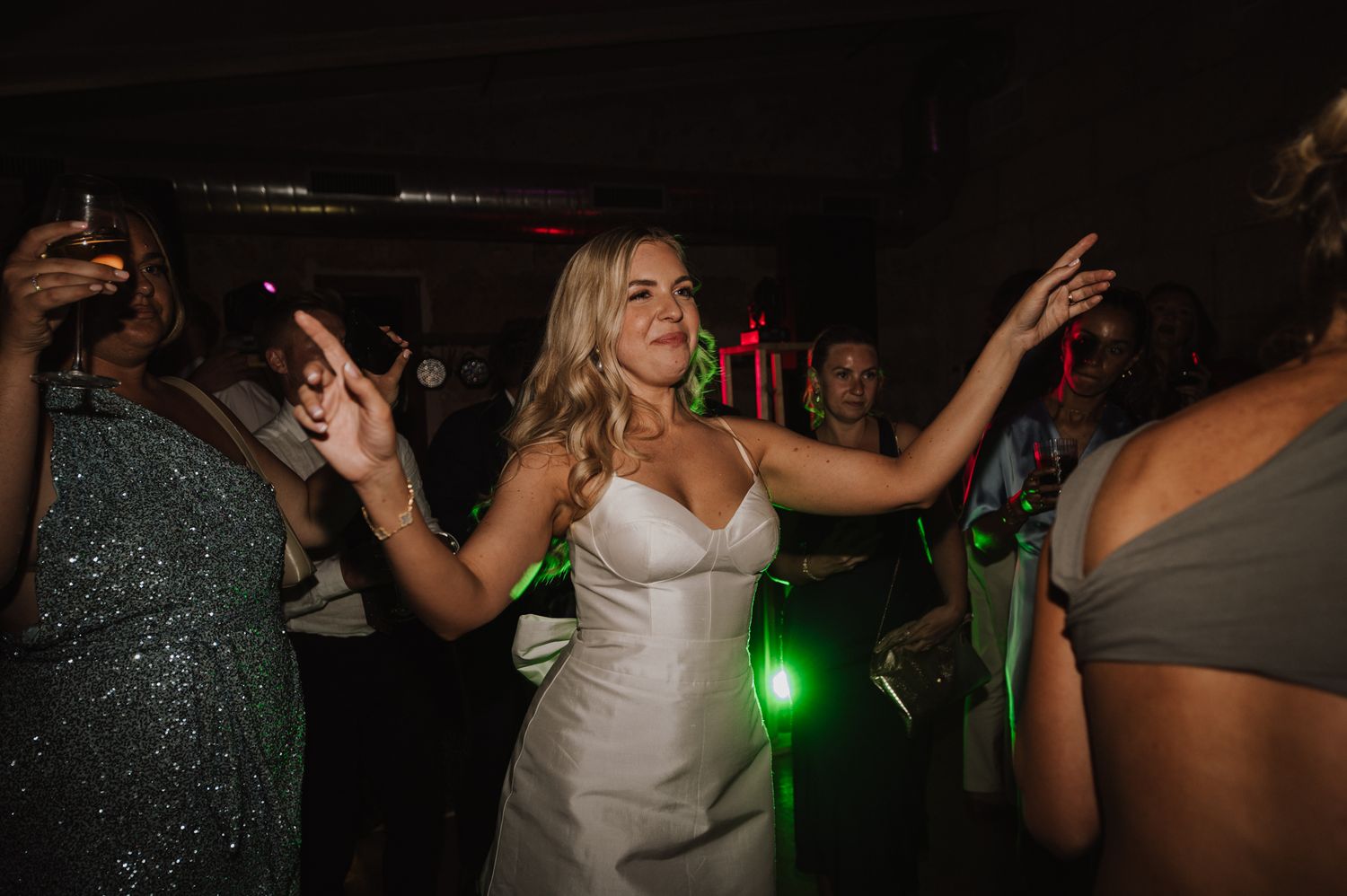 Bride in white gown dancing joyfully with guests under colorful lights at wedding reception party.