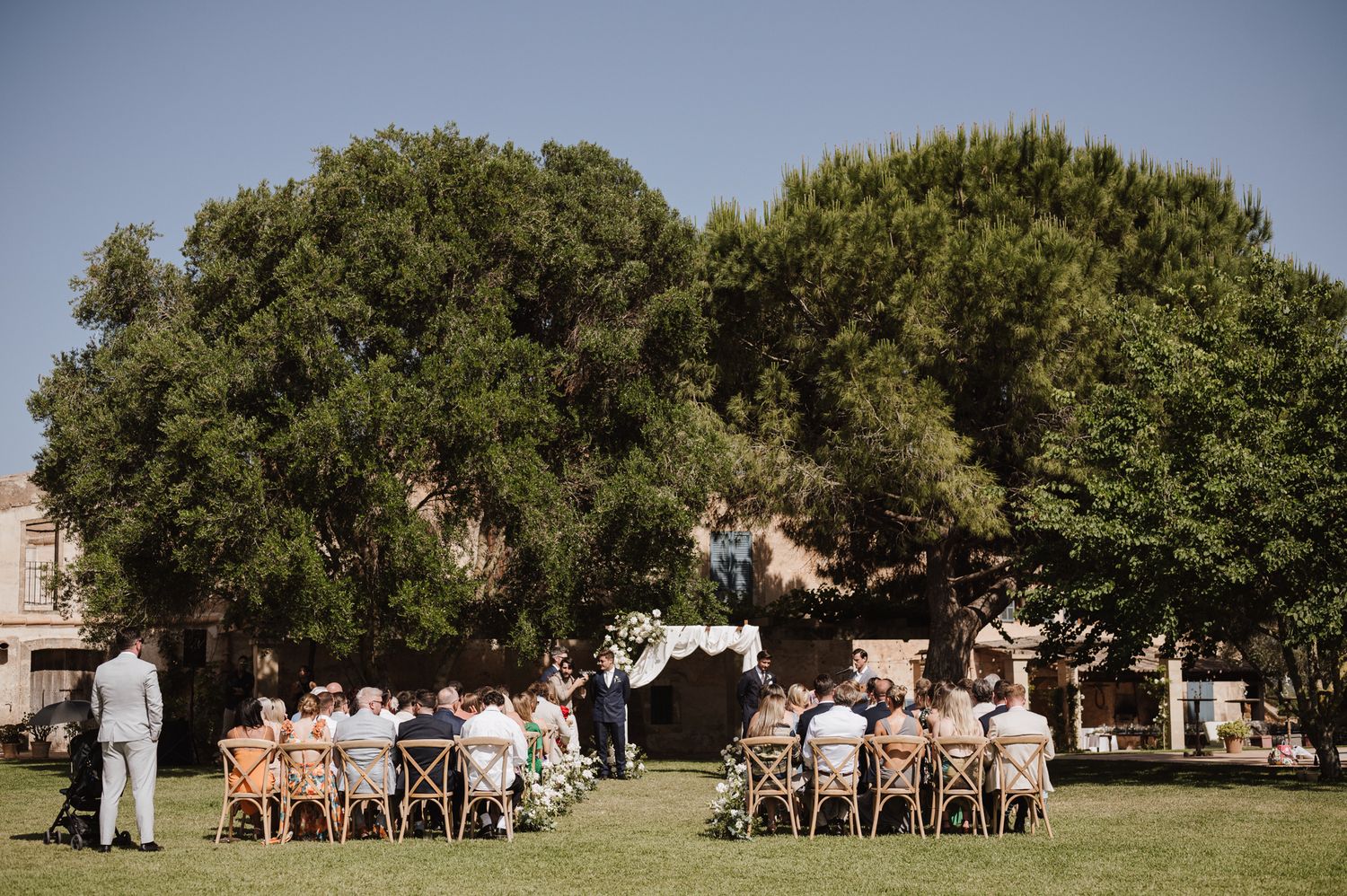 Intimate outdoor wedding ceremony with guests seated on wooden chairs beneath large trees at a rustic estate.