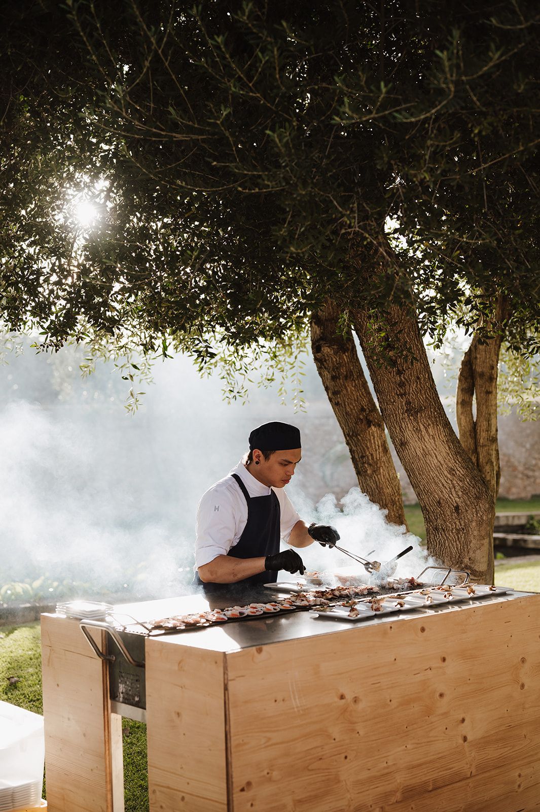 Chef grilling seafood skewers at outdoor wedding catering station with dramatic smoke rising.
