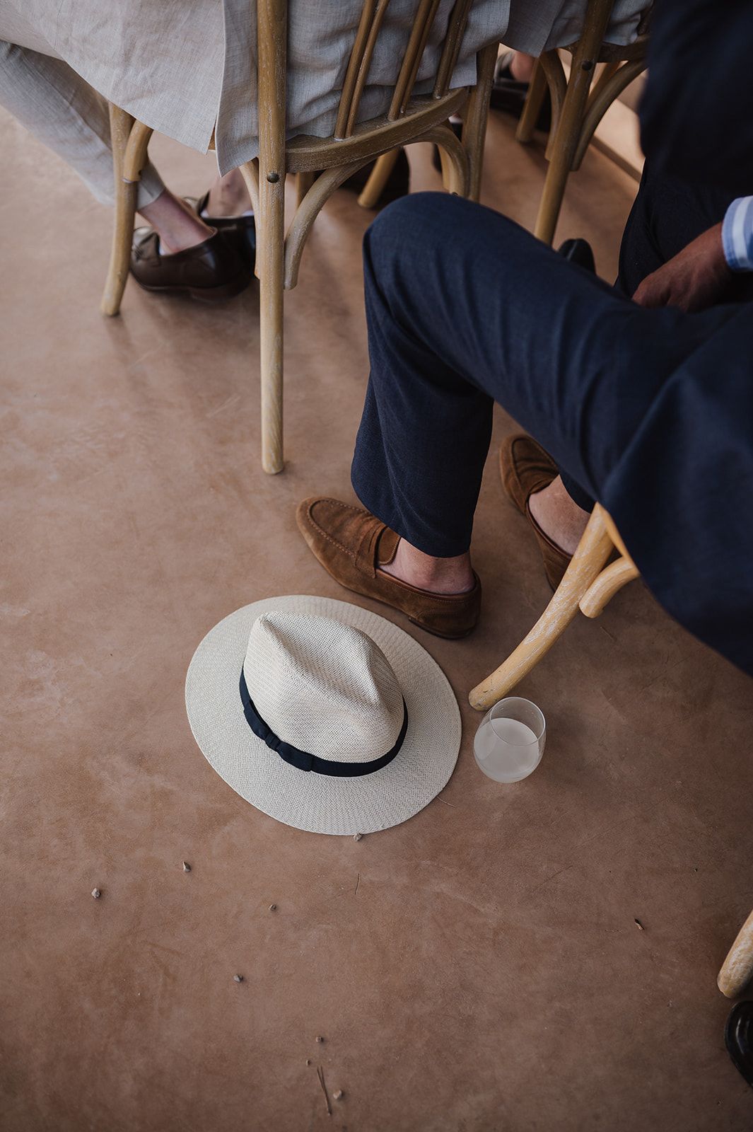 Groom in navy suit with brown loafers sits beside a white panama hat and glass on a concrete floor.