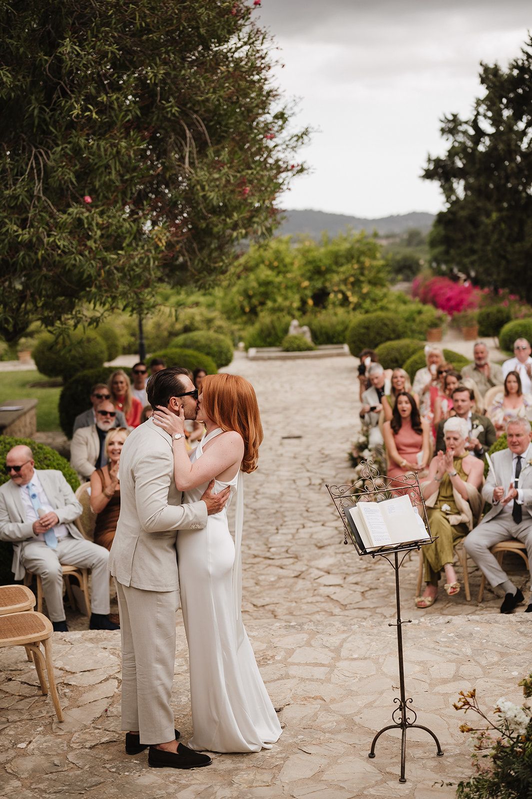 Newlyweds share first kiss as wedding guests cheer and applaud at outdoor garden ceremony.