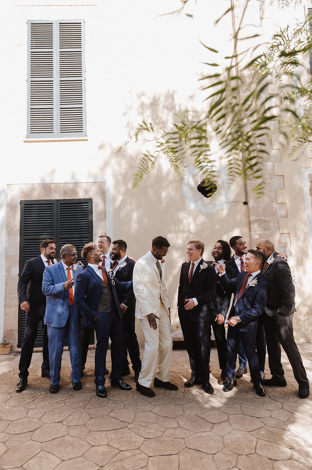 Groom in ivory suit celebrating with groomsmen in navy and blue suits at outdoor wedding venue.