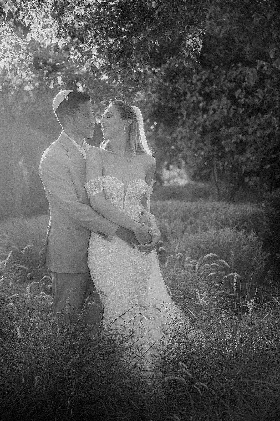 Black and white photo of bride and groom embracing in lush garden during golden hour wedding portrait.