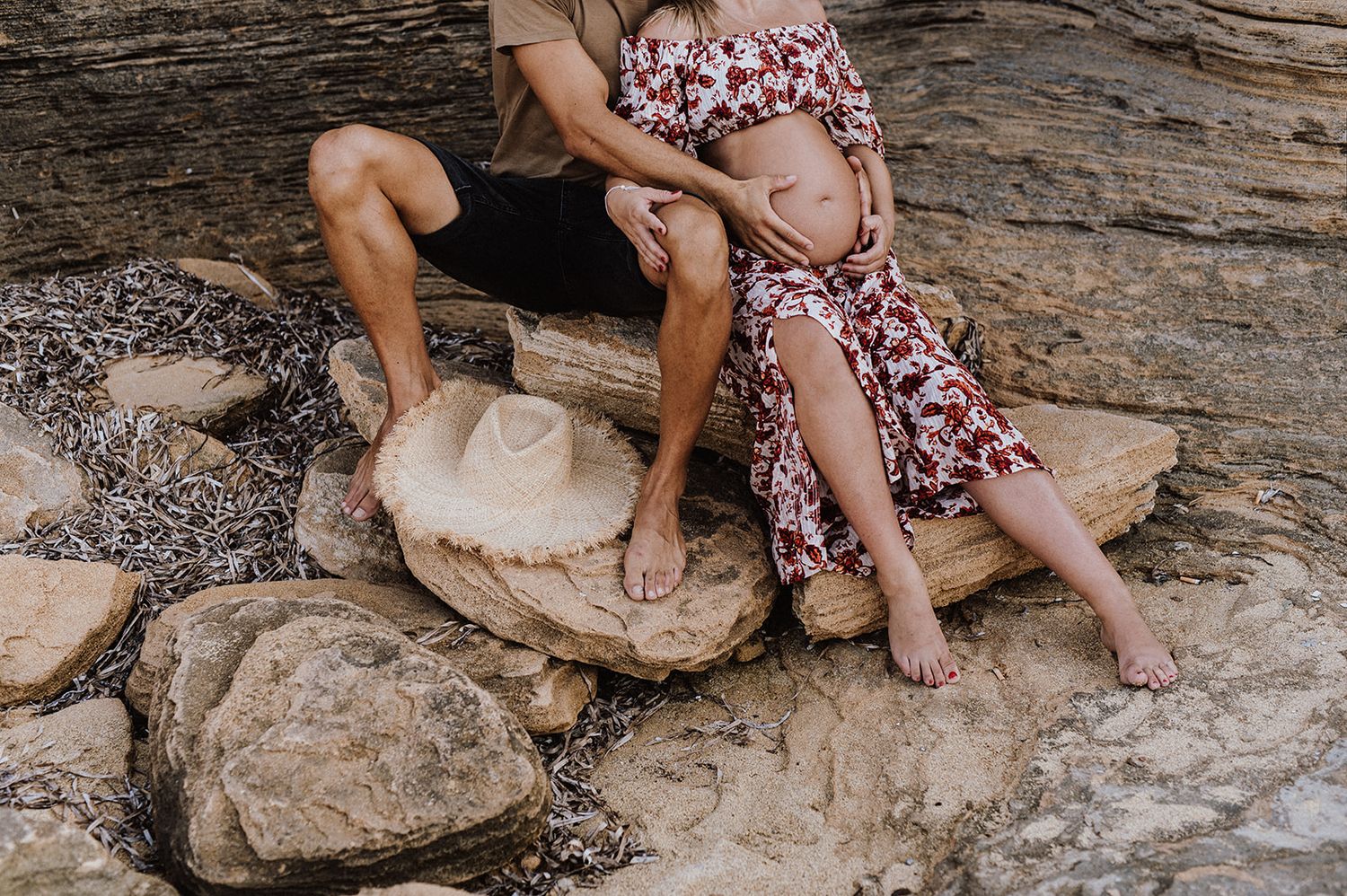 Couple sitting on rocks outdoors, woman in floral dress showing baby bump, man in shorts beside her with a hat on rocks.