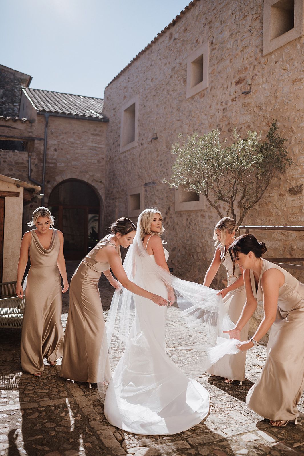 Bride in white gown and veil surrounded by bridesmaids in gold dresses in a rustic stone courtyard.
