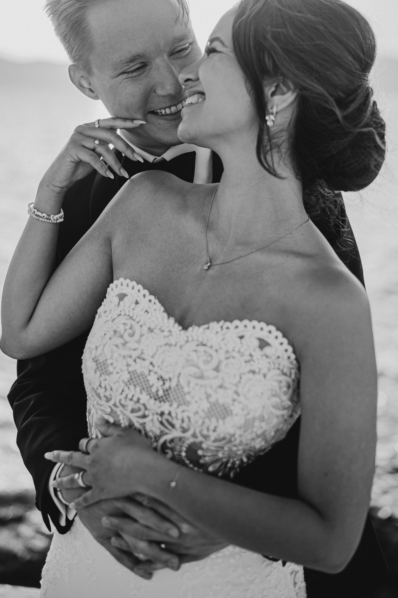 Black and white wedding photo of smiling bride in lace strapless gown embraced by groom in tuxedo outdoors.