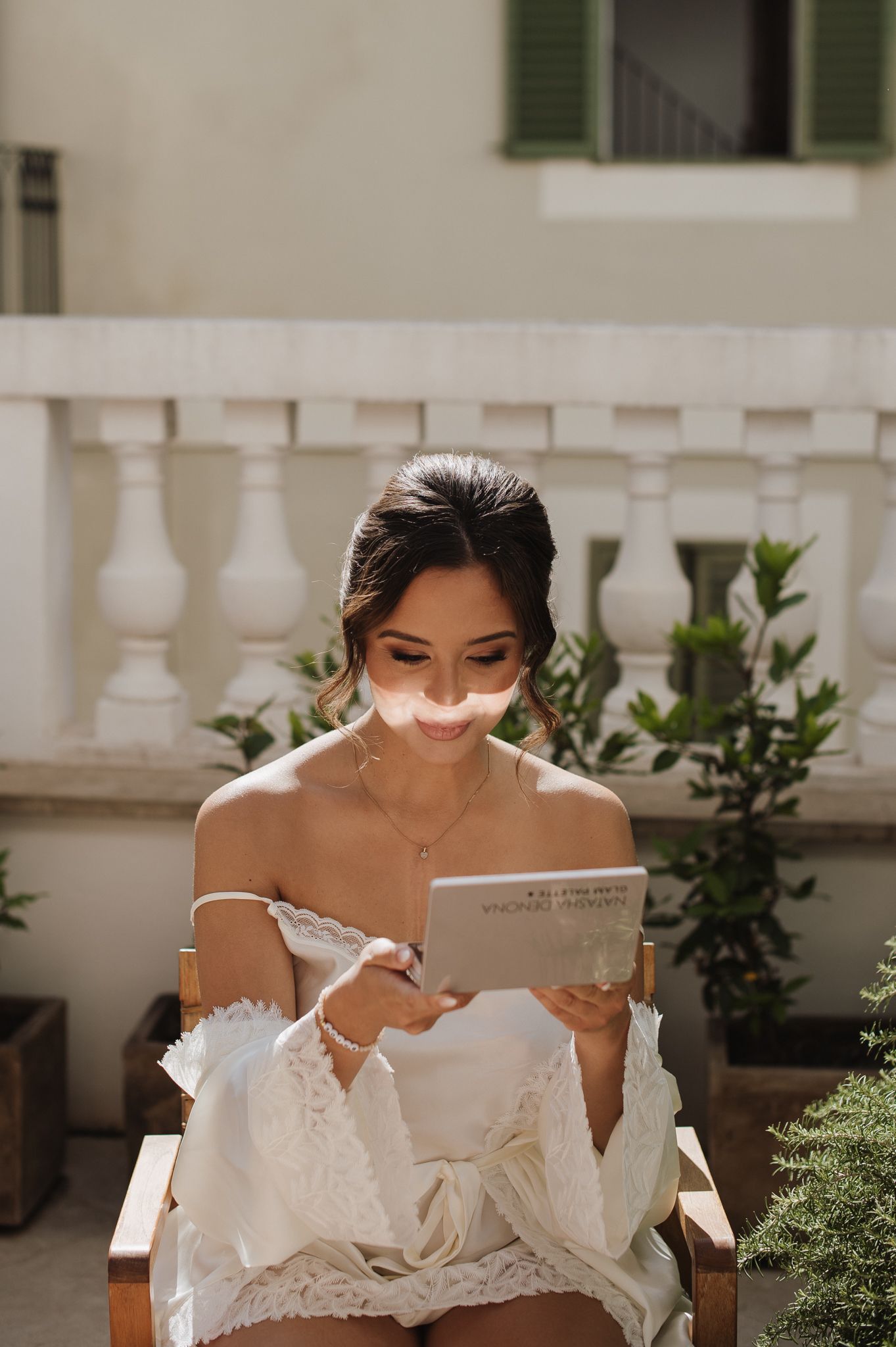 Bride in white off-shoulder lace dress reads a letter while seated on a chair on a sunlit balcony.