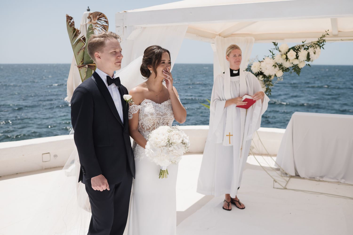 Bride and groom exchanging vows at oceanfront wedding ceremony with officiant and white floral arch backdrop.