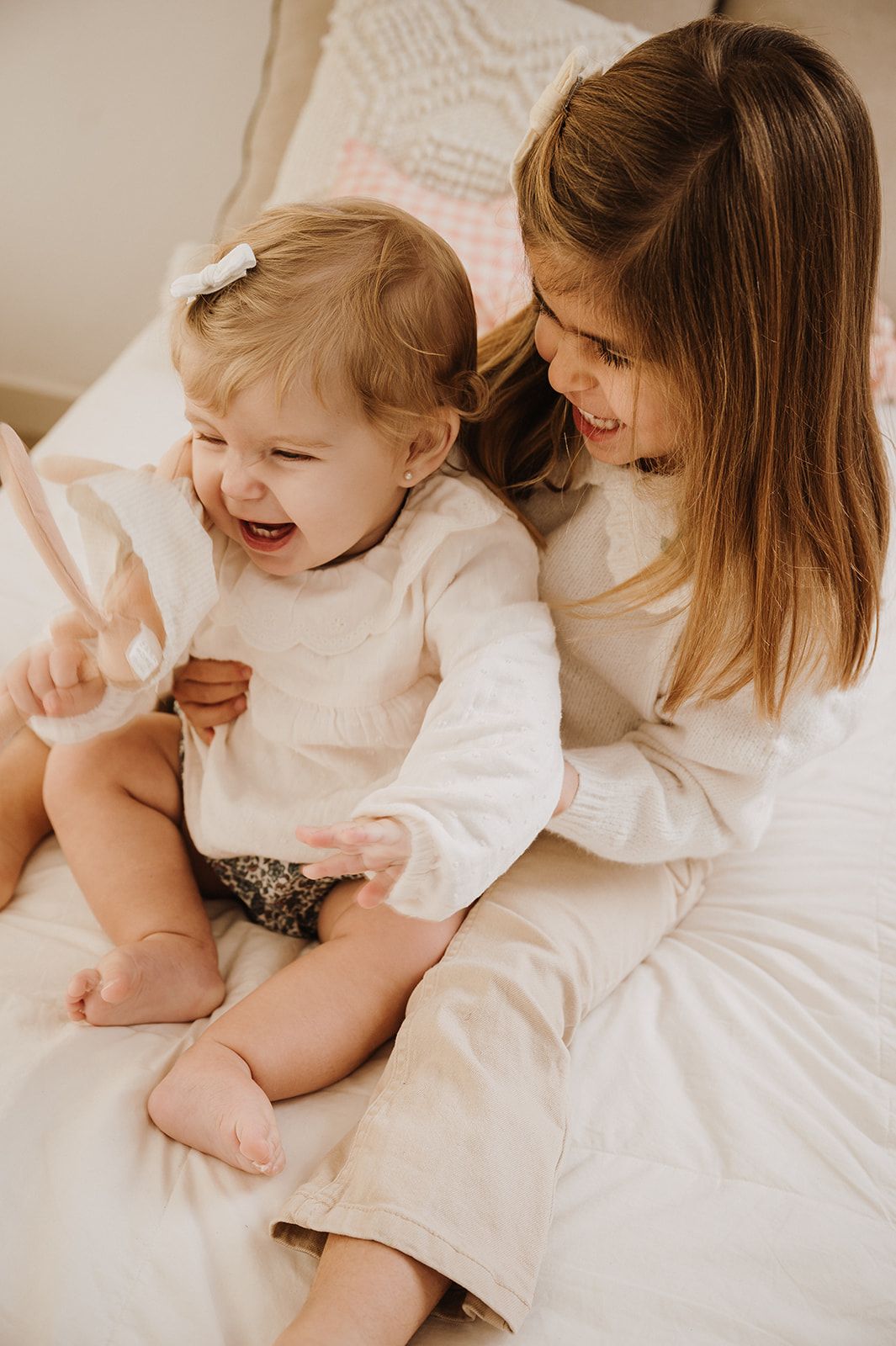 Mother sitting on bed smiling at laughing baby girl in white onesie during a joyful moment.