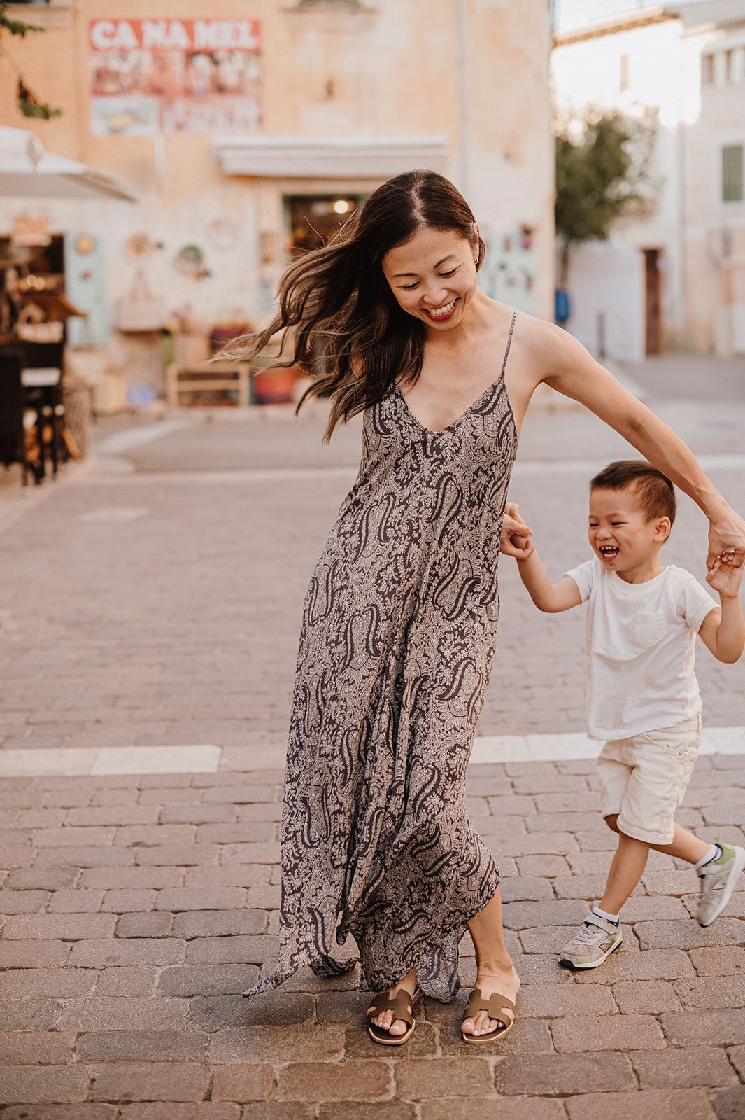 A joyful Asian mother in a paisley maxi dress holds hands with her laughing toddler son on a cobblestone street.