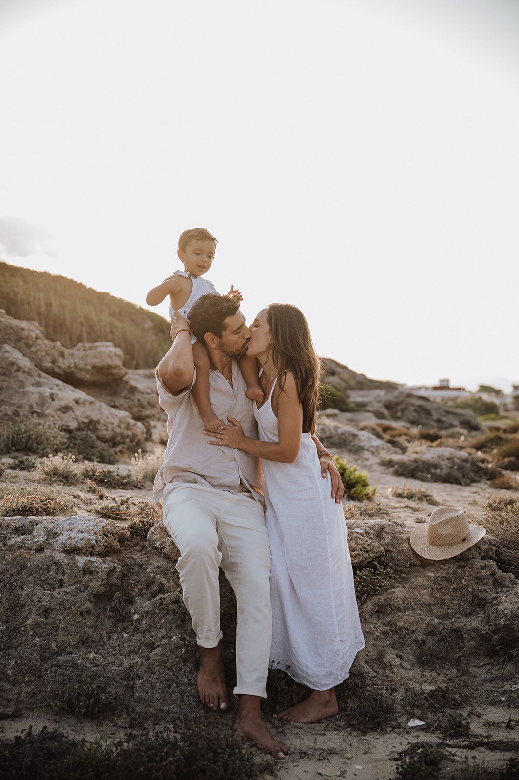 A couple shares a tender kiss while their toddler sits on dad's shoulders at a rocky sunset beach.