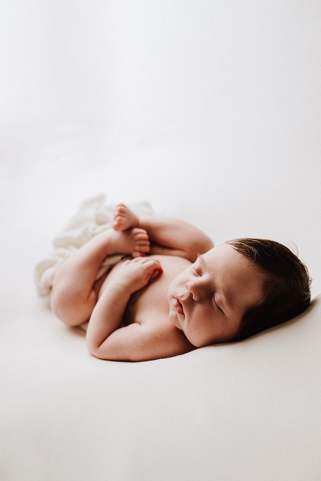 Peaceful newborn baby sleeping curled up on a white background, with dark hair and tiny hands tucked under chin.