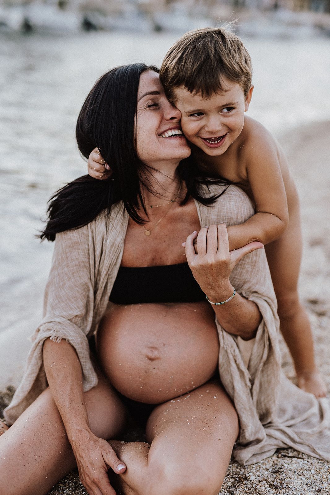 Pregnant woman sitting on beach smiling with young boy hugging her from behind near water.