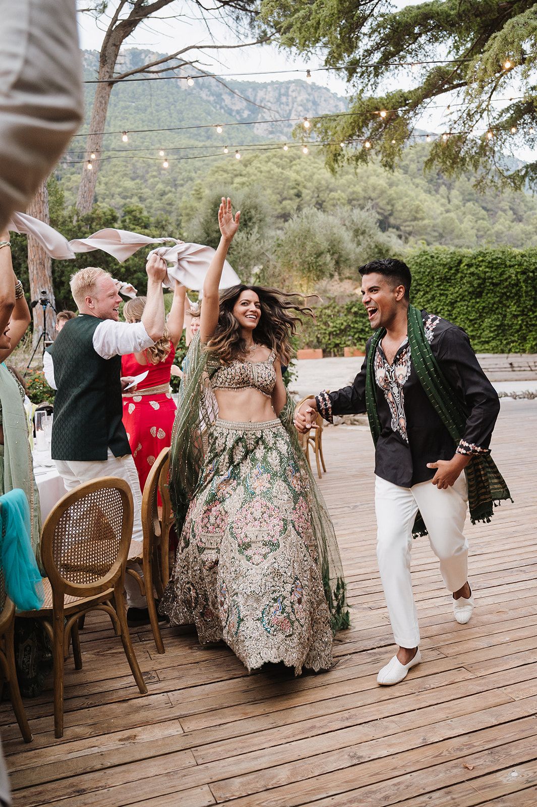 Couple making joyful entrance in Indian wedding attire at outdoor reception with string lights.