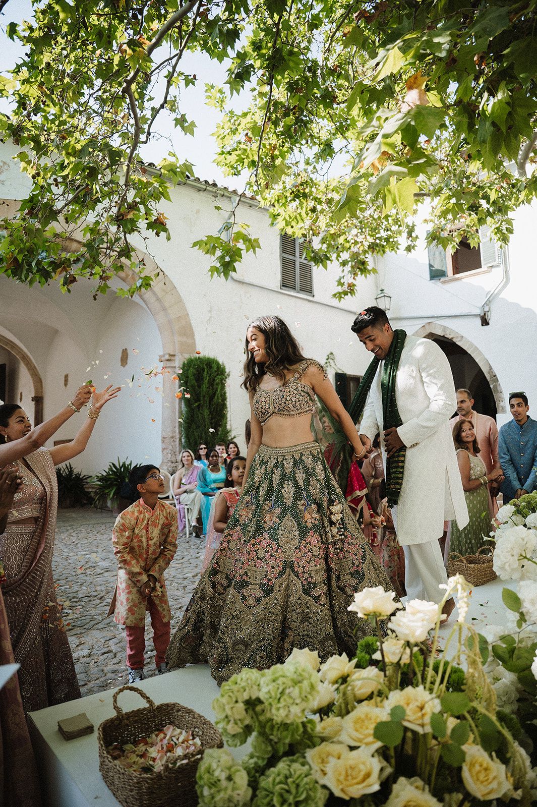 Bride in green lehenga descends steps with groom in white sherwani at outdoor Indian wedding ceremony.