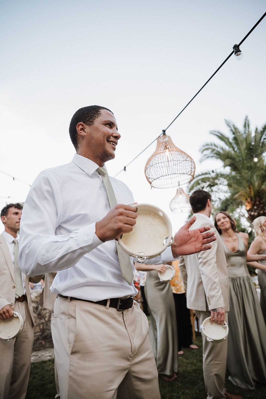 Smiling groomsman holds a tambourine during a lively outdoor wedding celebration under string lights.