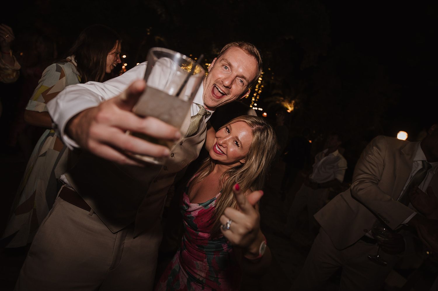 Wedding guests celebrate joyfully at lively nighttime reception, raising drinks and smiling at the camera.