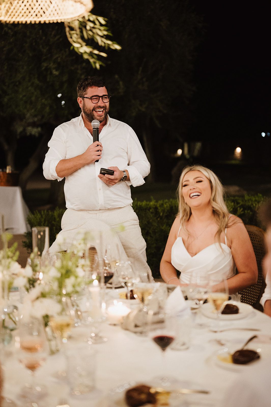 Man in white shirt gives a wedding speech into microphone as blonde bride laughs joyfully at candlelit table.