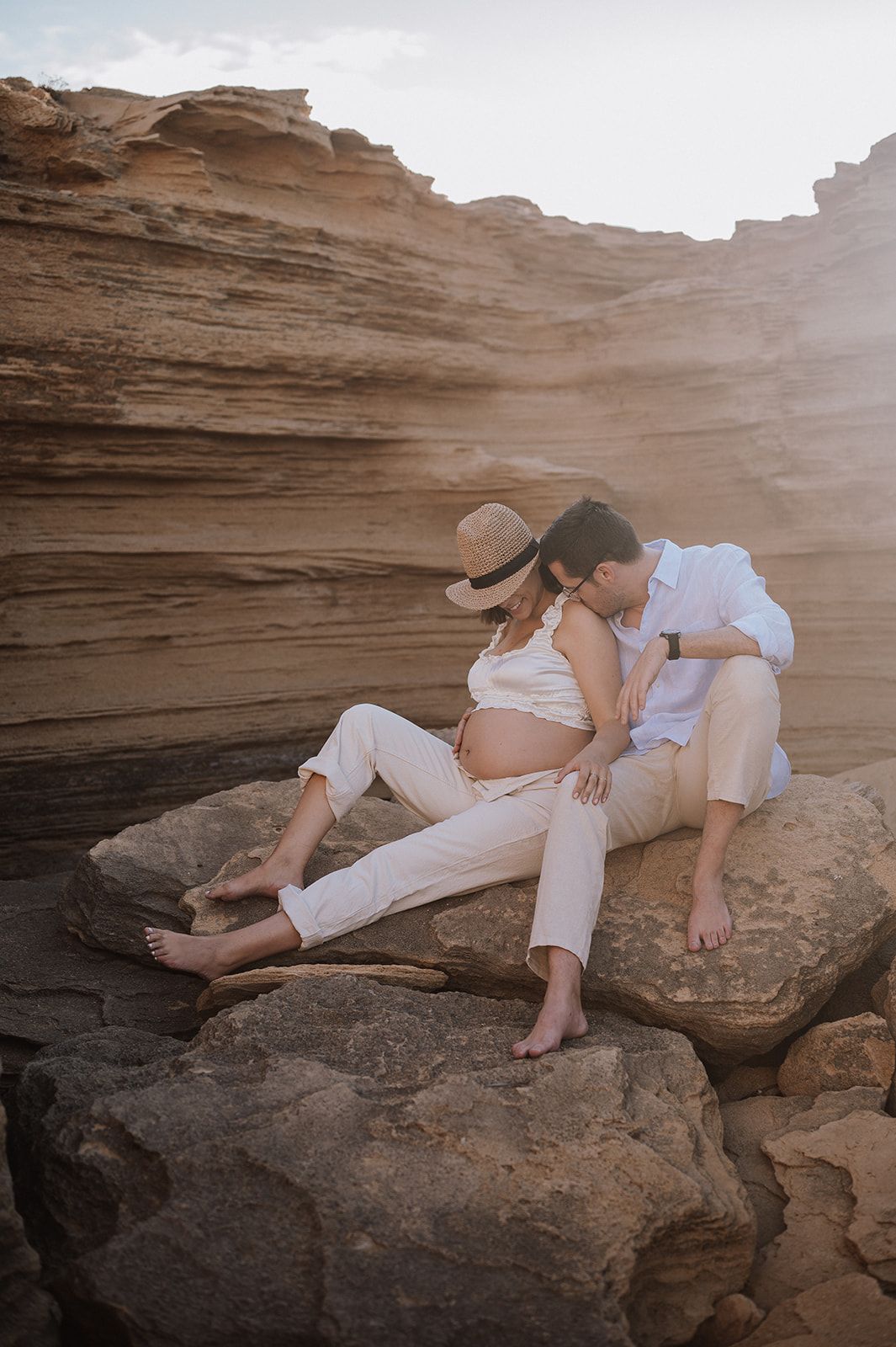 Pregnant couple sitting on rocky cliffs, woman in hat and white outfit, man kissing her shoulder tenderly.