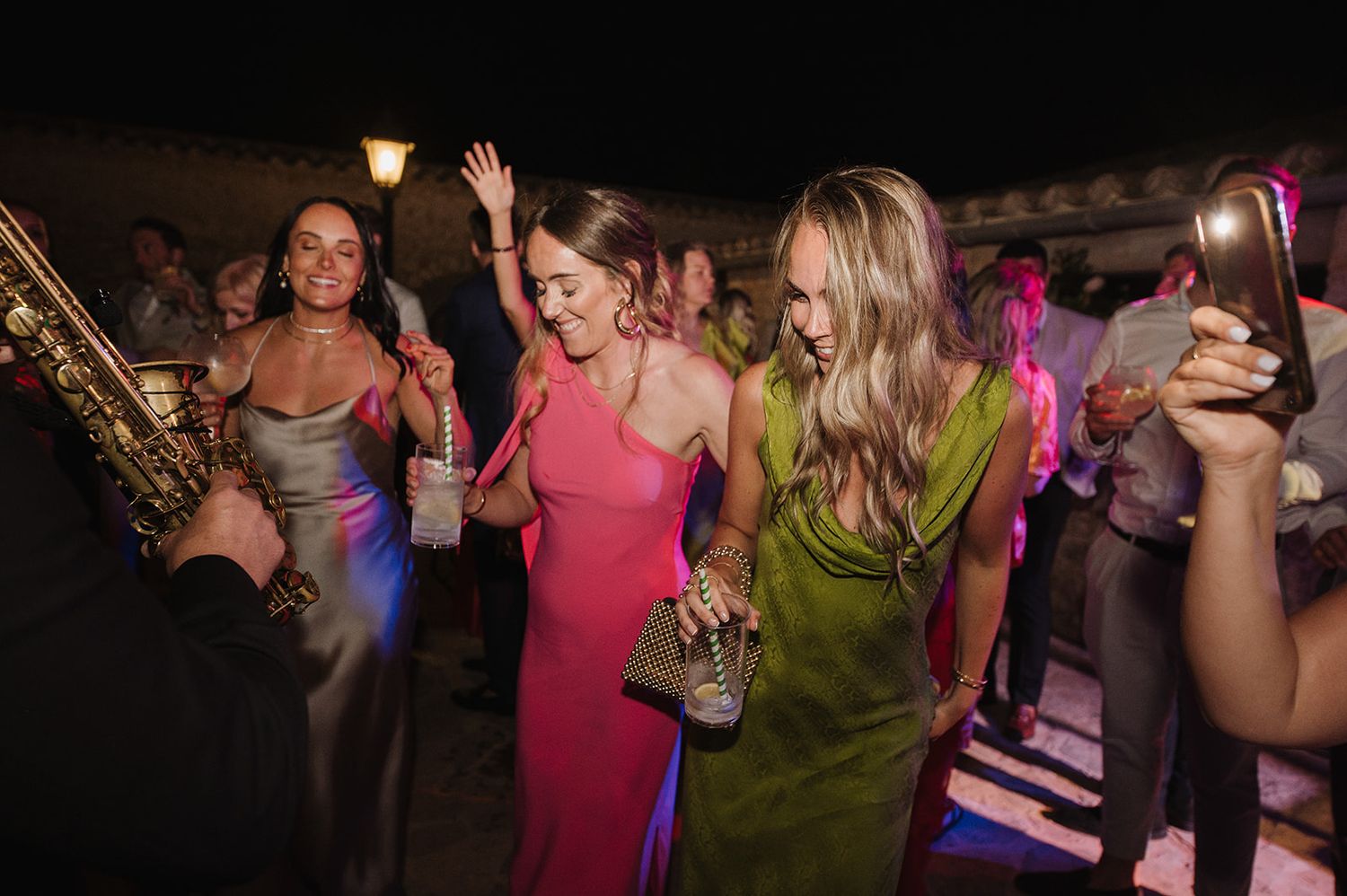 Women in colorful dresses dancing outdoors at a festive evening wedding reception.