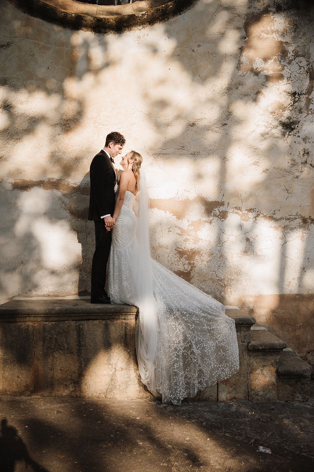 Bride and groom share intimate moment on stone steps with flowing lace veil against rustic wall.