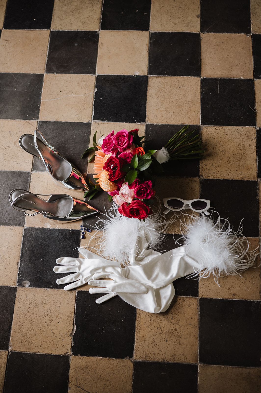 Bridal accessories flat lay with red bouquet, silver heels, white gloves and feathers on checkered floor.