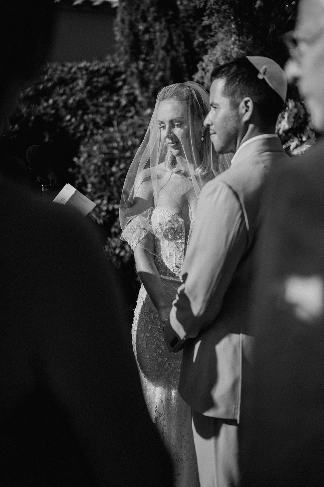 Black and white photo of smiling bride in lace gown and groom in kippah during outdoor ceremony.