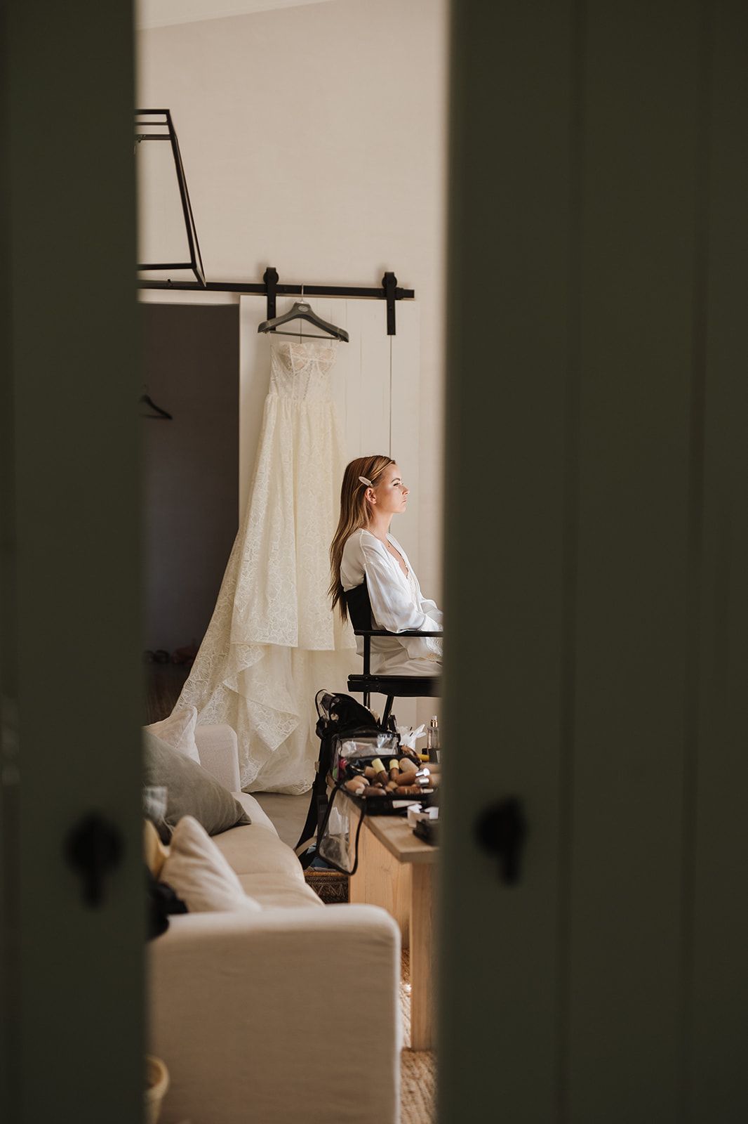 Bride in white robe sits before her wedding gown hanging in a softly lit bridal suite.