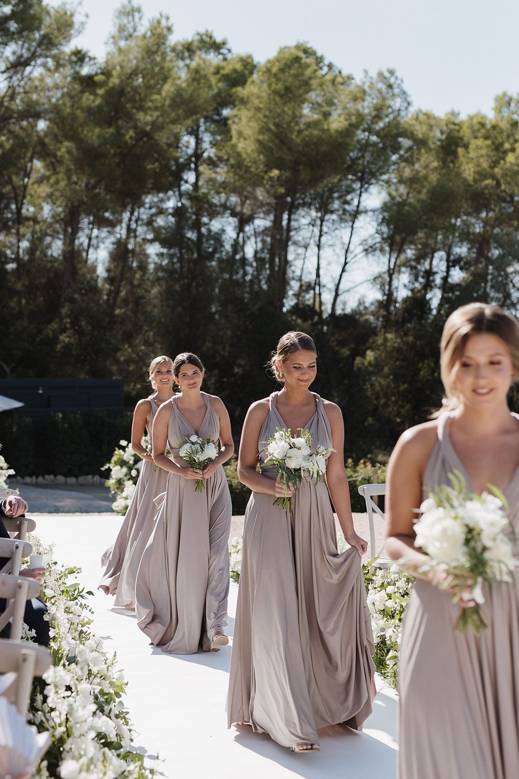Bridesmaids in elegant taupe gowns walk down the aisle carrying white floral bouquets at an outdoor wedding.