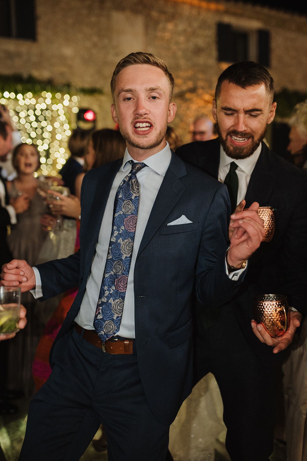 Two men in suits dancing and celebrating at an outdoor evening wedding reception with string lights.