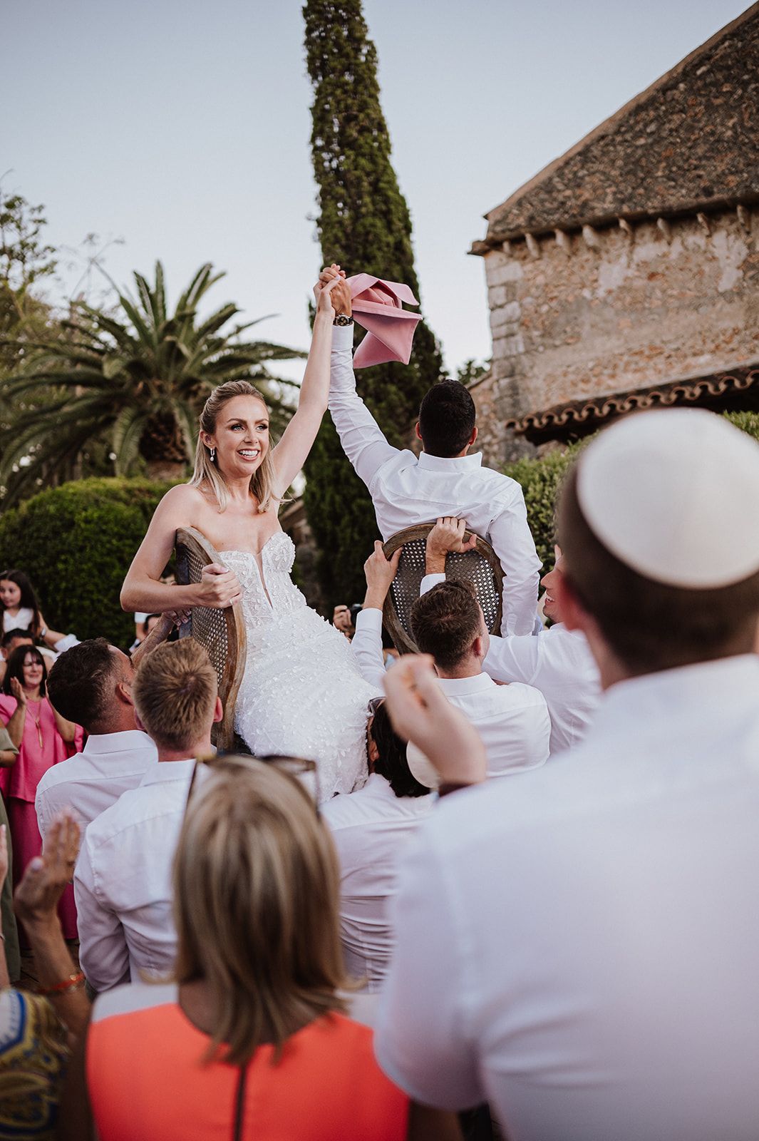 Bride and groom lifted in chairs during traditional Jewish Hora dance at outdoor wedding celebration.