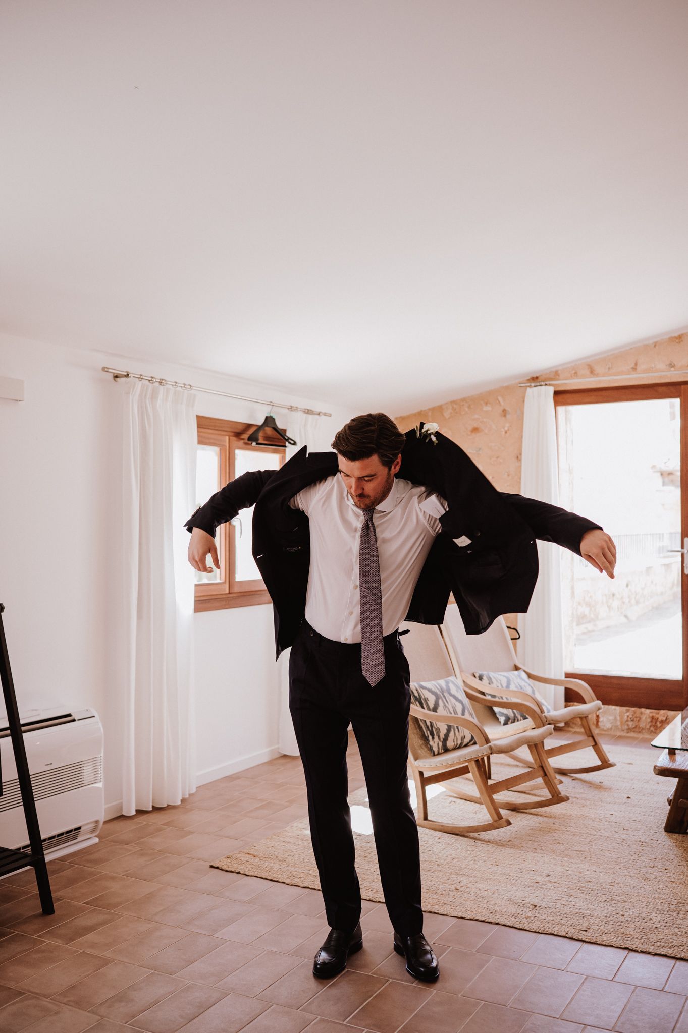Groom putting on his dark suit jacket while getting ready indoors before his wedding ceremony.