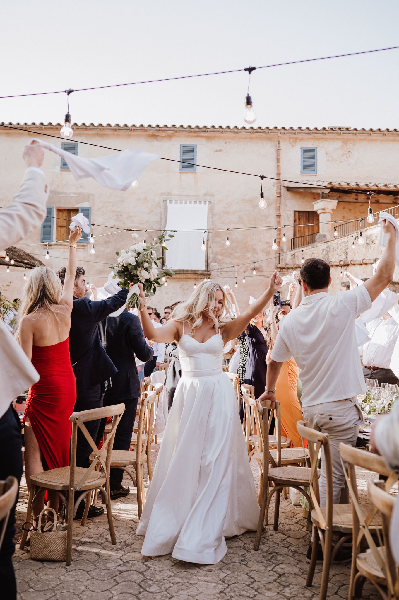 Jubilant bride walking down aisle as guests wave white handkerchiefs at rustic outdoor wedding ceremony.
